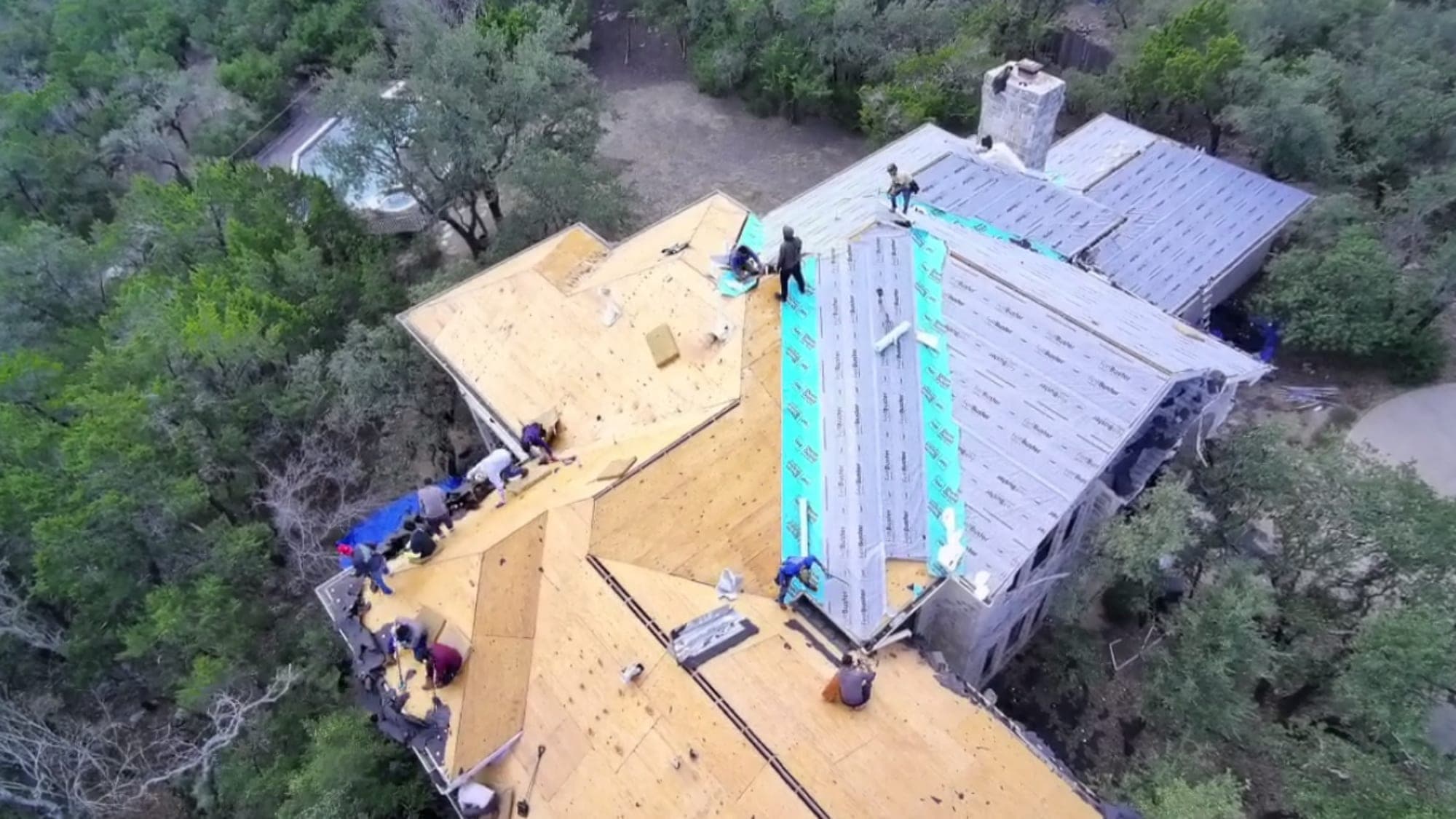 Overhead drone view of a complex multi-plane roof, fresh decking on one side and synthetic underlayment on the other, pool below