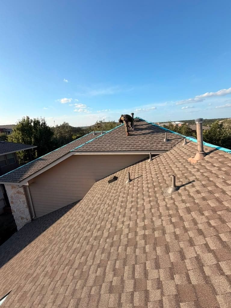 Crew member walking a long finished ridge on a tan architectural shingle roof under blue sky
