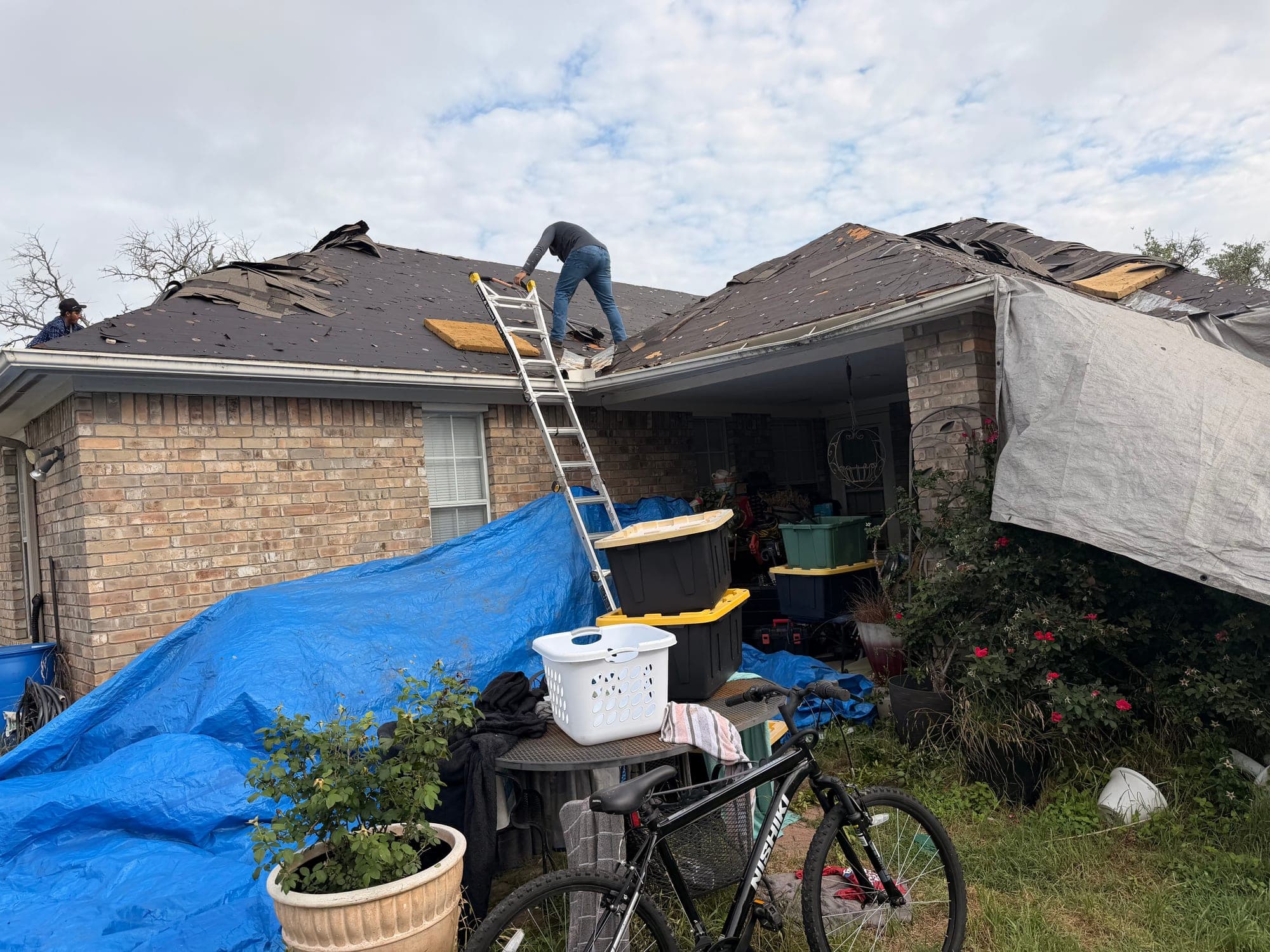 Storm damaged brick home with shingles peeled back, tarps covering the patio and the homeowner's belongings